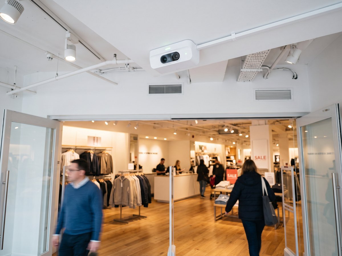 People counter sensor mounted on the ceiling above a store entrance with shoppers walking by